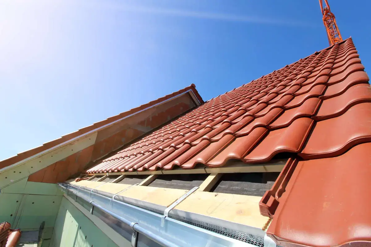 Taktegel i rött läggs på ett hus under uppbyggnad. En kran syns i bakgrunden, redo att lyfta material för framtida byggnadsintegrerade solceller.