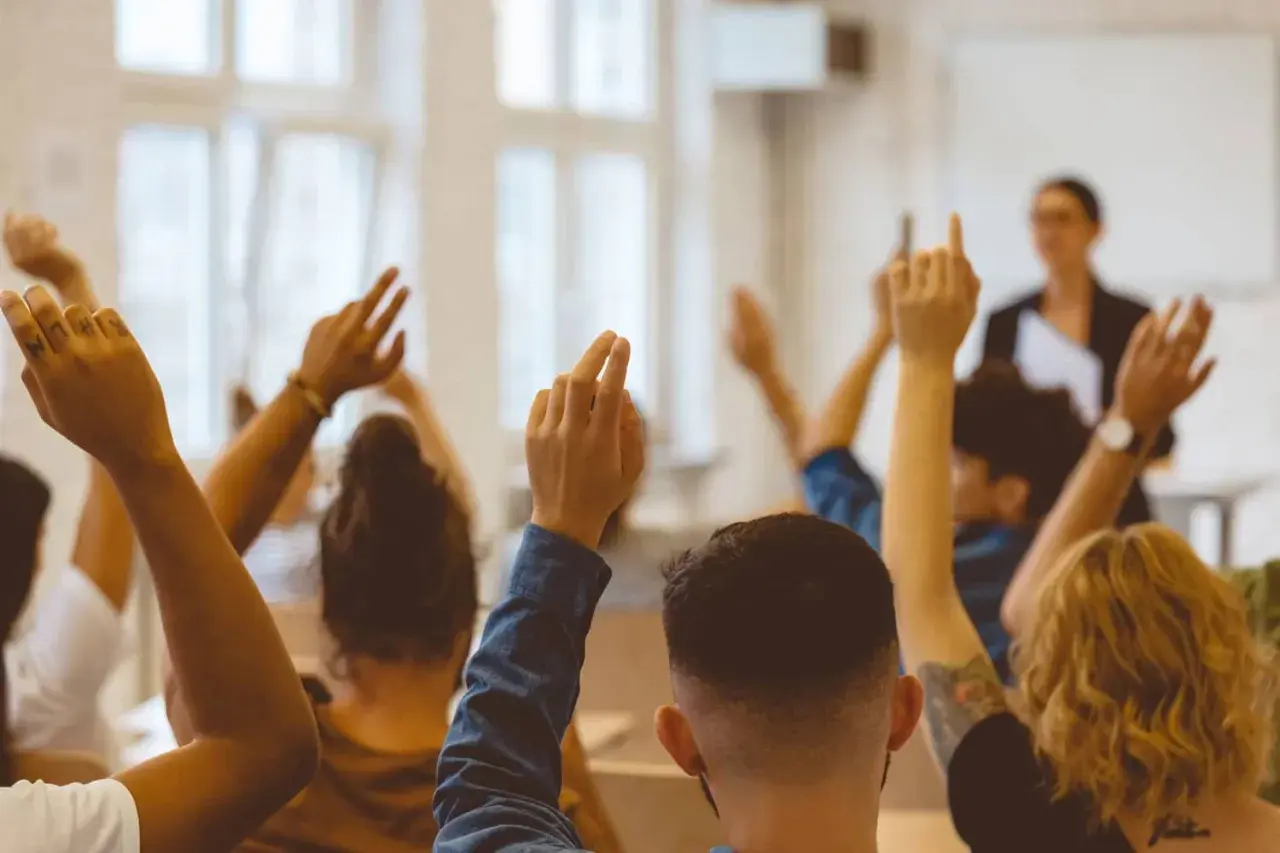 Schüler heben die Hände, um Fragen zu stellen. Ein achtsamkeit lehrer im Hintergrund beobachtet die Klasse.