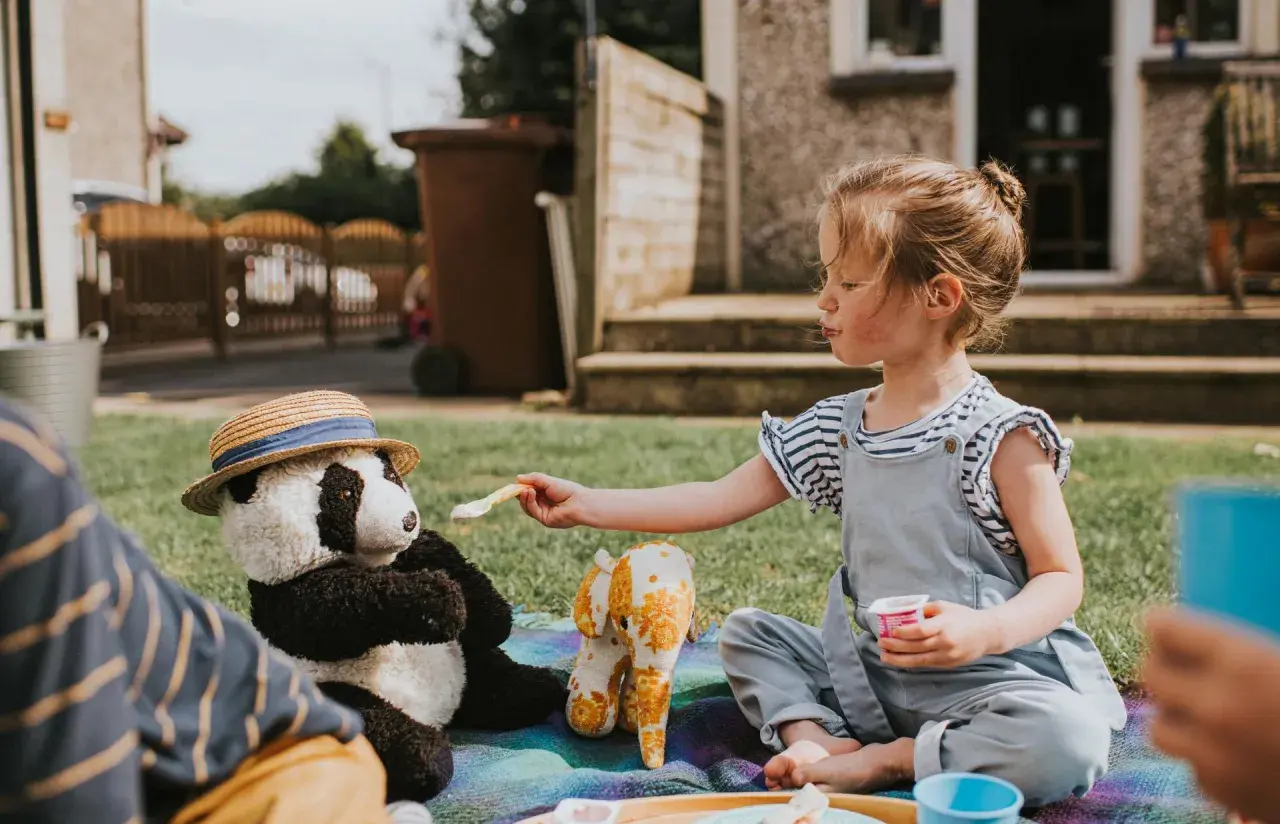 Dziewczynka karmi pluszową pandę w kapeluszu na pikniku na trawie.