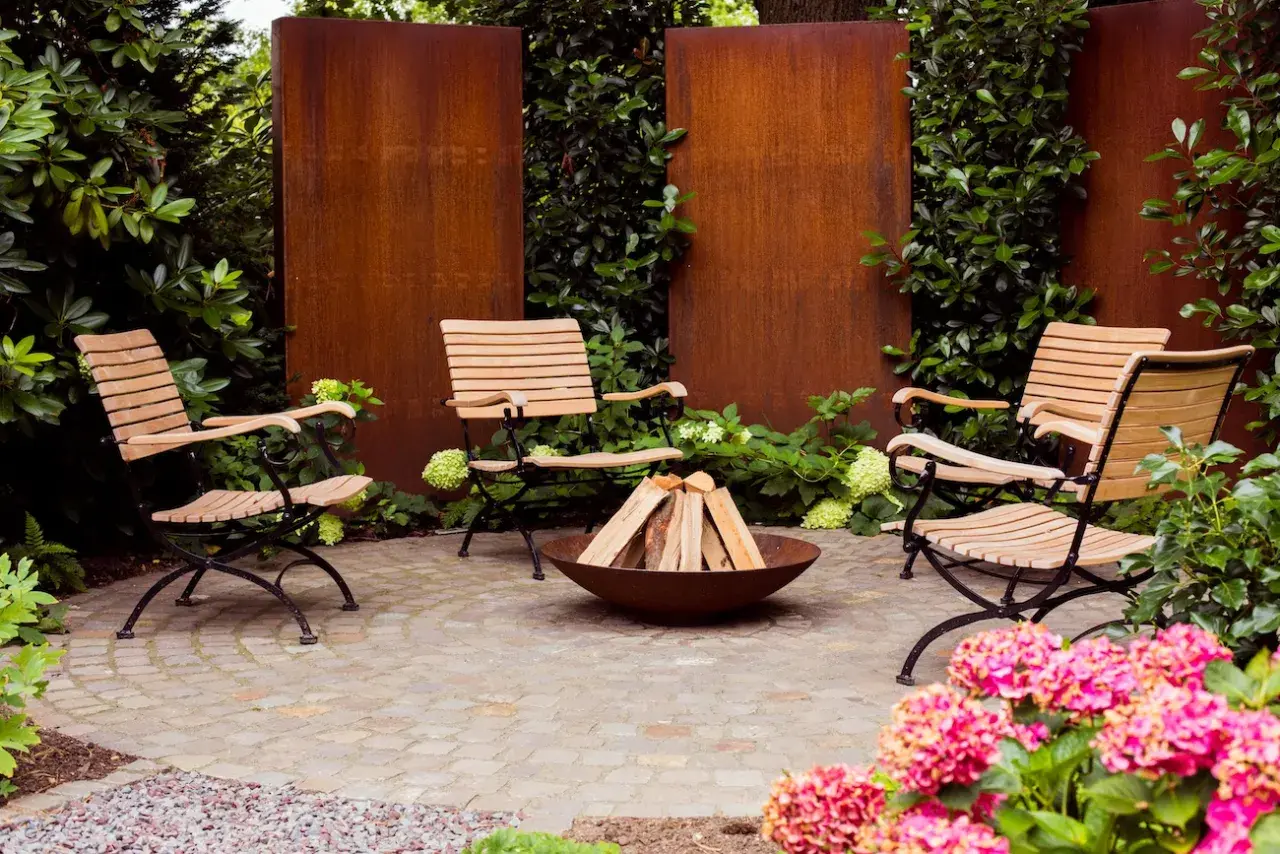 Moderne, schlichte Gartengestaltung mit vier Holzstühlen um eine Feuerstelle. Rostige Metallwände und üppiges Grün schaffen eine gemütliche Atmosphäre.