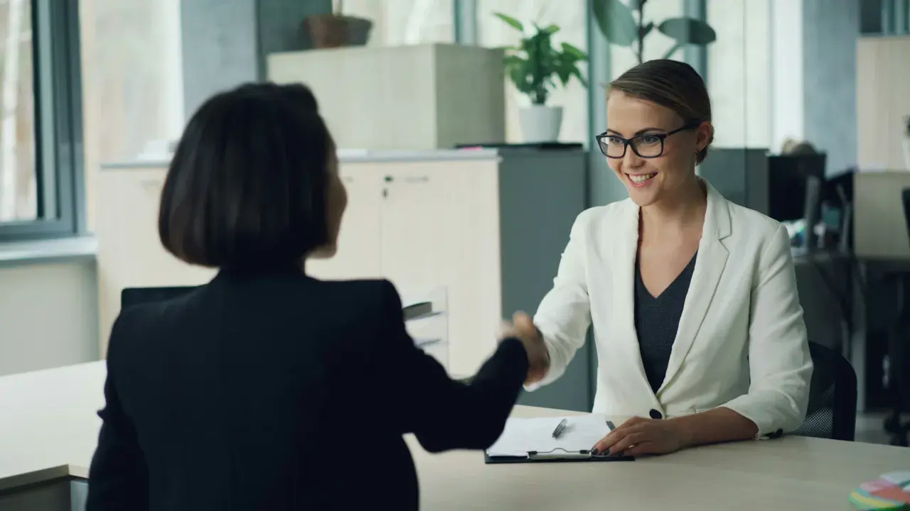 Zwei Frauen schütteln sich die Hände. Eine Frau im weißen Blazer lächelt, während sie eine Bewerbung für personalentwicklung jobs prüft.