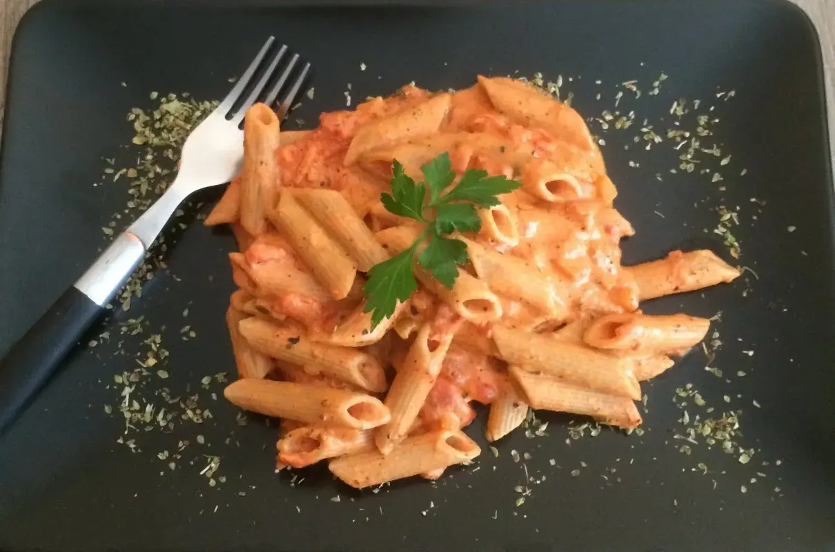 Penne w kremowym sosie pomidorowym z mascarpone, udekorowane natką pietruszki. Obok widelec i posypka z ziół.