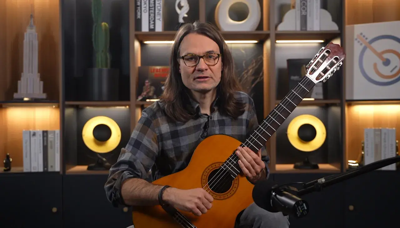 Mężczyzna z gitarą uczy, jak nauczyć się nut. W tle półki z książkami i dekoracjami.