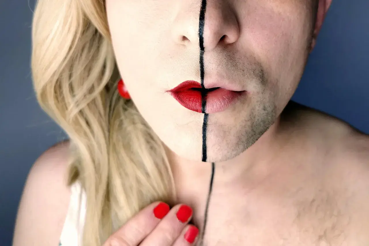 Un volto diviso a metà, con capelli biondi e barba, labbra rosse e una linea nera. Un'espressione di una persona non binaria che abbraccia la propria identità.