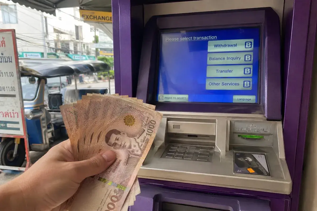 Hand hält thailändische Baht-Scheine vor einem Geldautomaten. Hier kann man Geld wechseln.