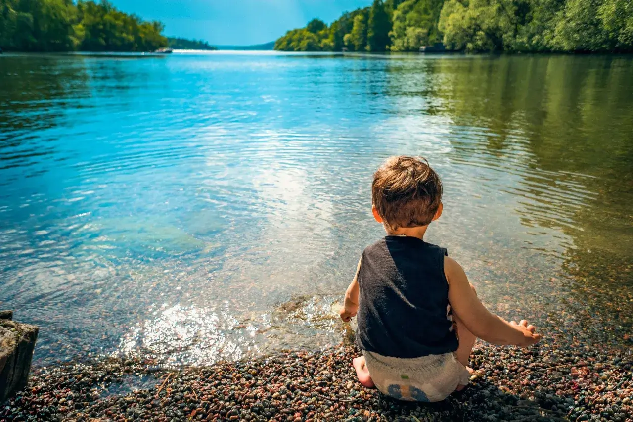Un bambino seduto sulla riva di un lago, godendosi un tranquillo week end con bambini. L'acqua limpida riflette il cielo azzurro e gli alberi verdi.