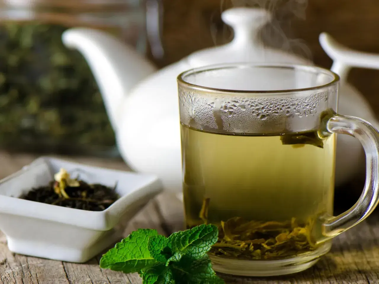 Dampfender grüner Tee in einer Tasse, daneben eine Schale mit Teeblättern und Minze. Welcher Tee ist antibakteriell? Grüner Tee ist bekannt für seine gesundheitlichen Vorteile.