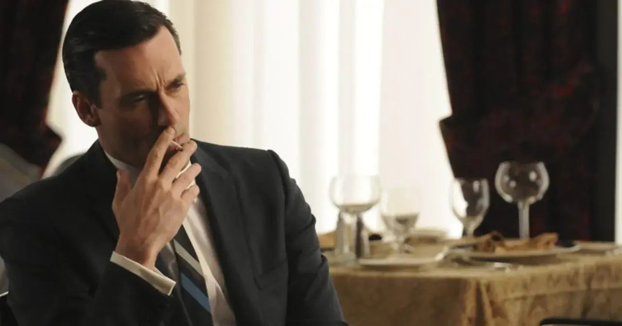 Jon Hamm, znany z filmów, seriali i programów, w eleganckim garniturze pali papierosa przy stole.