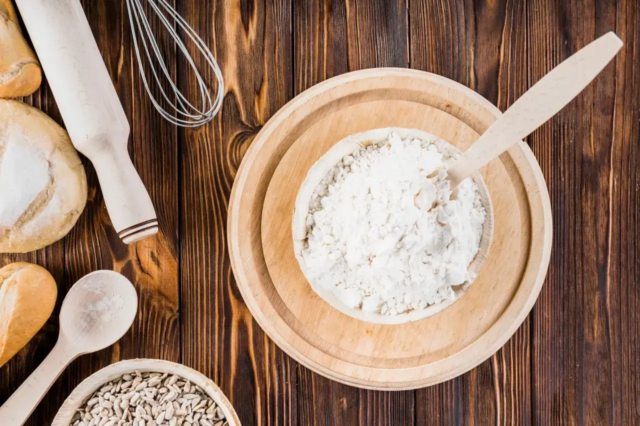 Mąka orkiszowa, podobnie jak pszenna, zawiera gluten. Na zdjęciu widać składniki do pieczenia: mąkę, chleb, ziarna słonecznika, wałek i trzepaczkę.