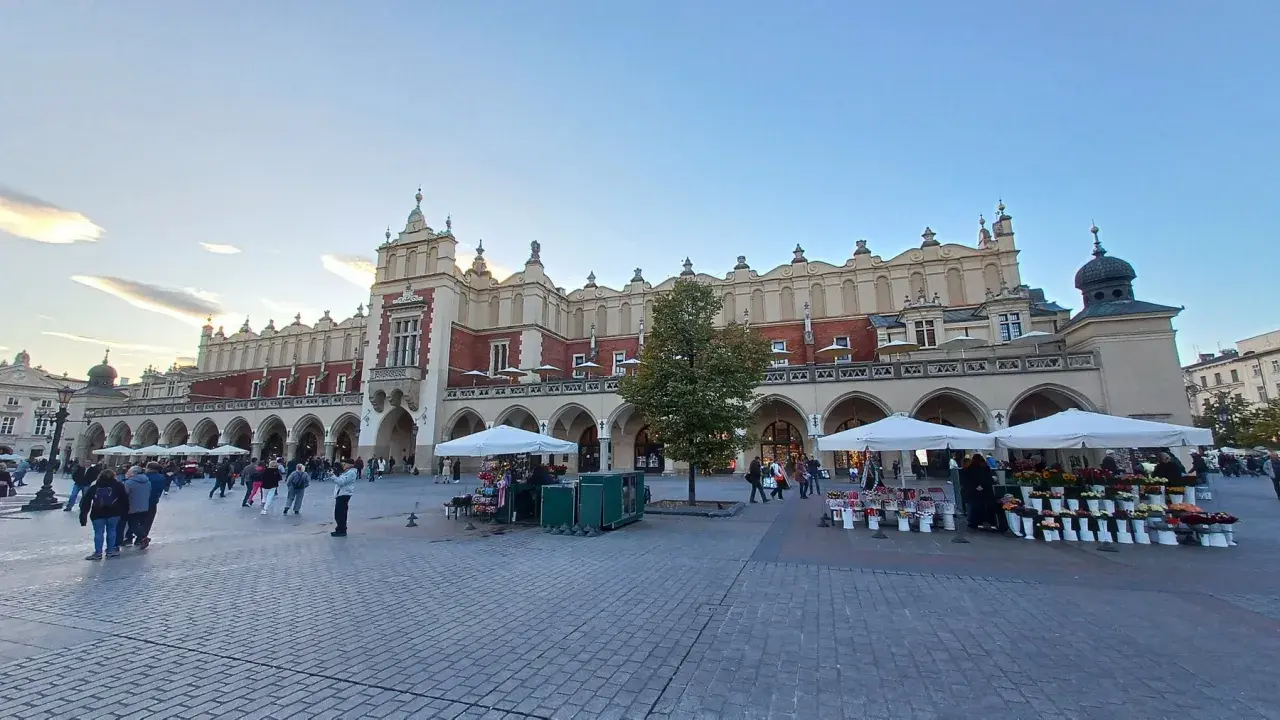 Sukiennice, jeden z najpiękniejszych zabytków w Krakowie, tętni życiem. Ludzie spacerują po Rynku Głównym, podziwiając architekturę i kupując kwiaty.