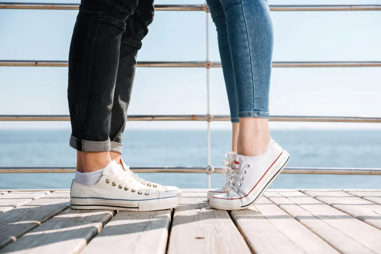 Para na drewnianym pomoście, w białych trampkach i jeansach. Idealne converse stylizacje damskie na letni dzień nad morzem.