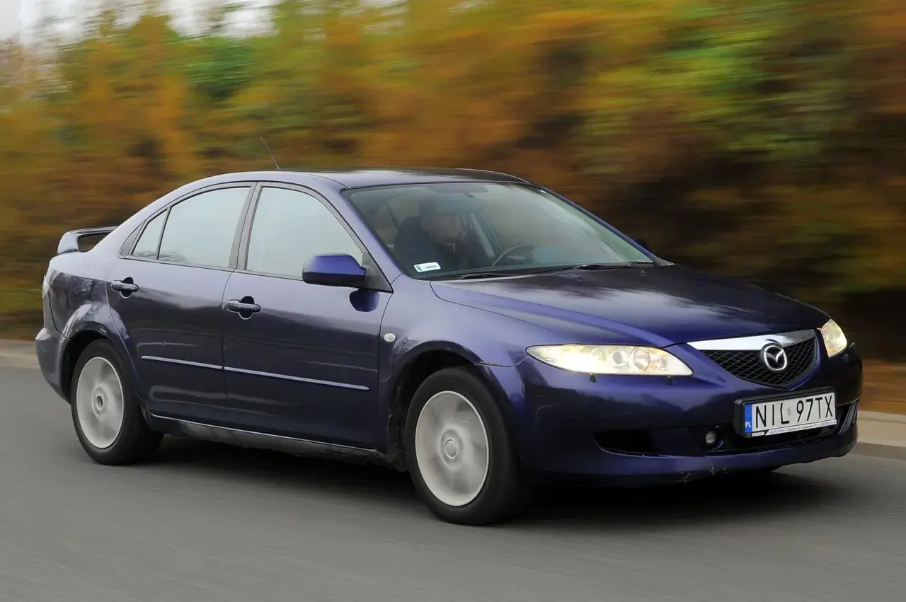 Granatowa Mazda 6 GG jedzie po drodze, za nią rozmyty krajobraz jesiennych drzew.