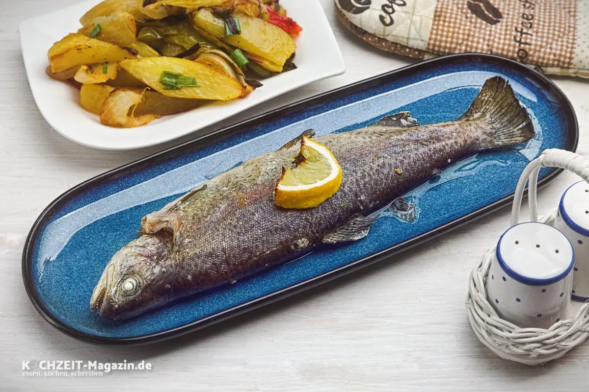 Ein ganzer, im Ofen gebackener Fisch auf einem blauen Teller, garniert mit einer Zitronenspalte. Daneben eine Portion Kartoffel-Gemüse-Spalten.