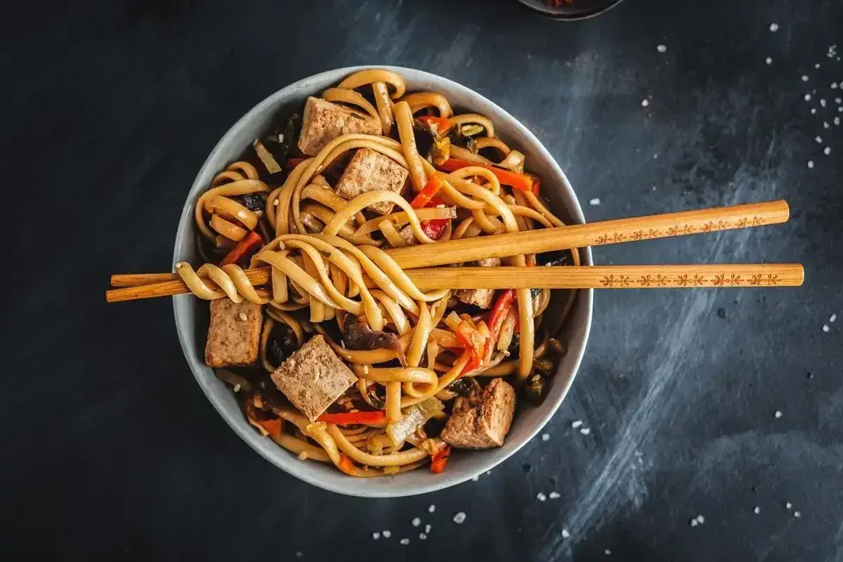 Miska z makaronem udon, tofu i warzywami, przykryta pałeczkami.