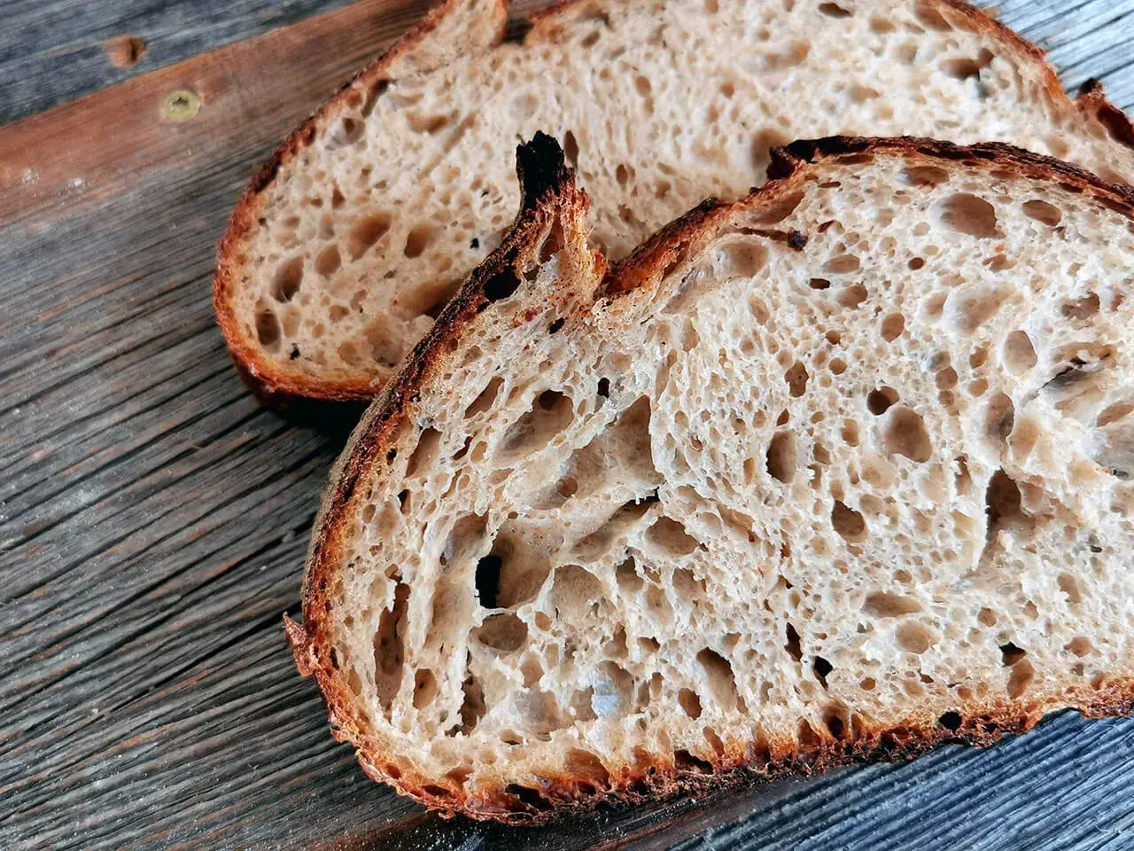 Zwei Scheiben rustikales Sauerteigbrot mit großer Porung liegen auf einem Holzbrett. Warum ist Sauerteig gesund? Weil die Fermentation Nährstoffe besser verfügbar macht.