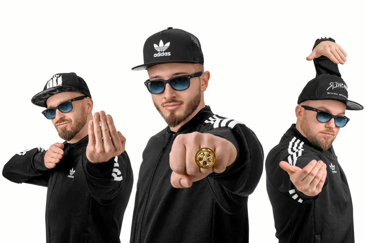 Trzech mężczyzn w czarnych bluzach z paskami Adidas i czapkach z daszkiem, pozujących jak zespoły disco polo.