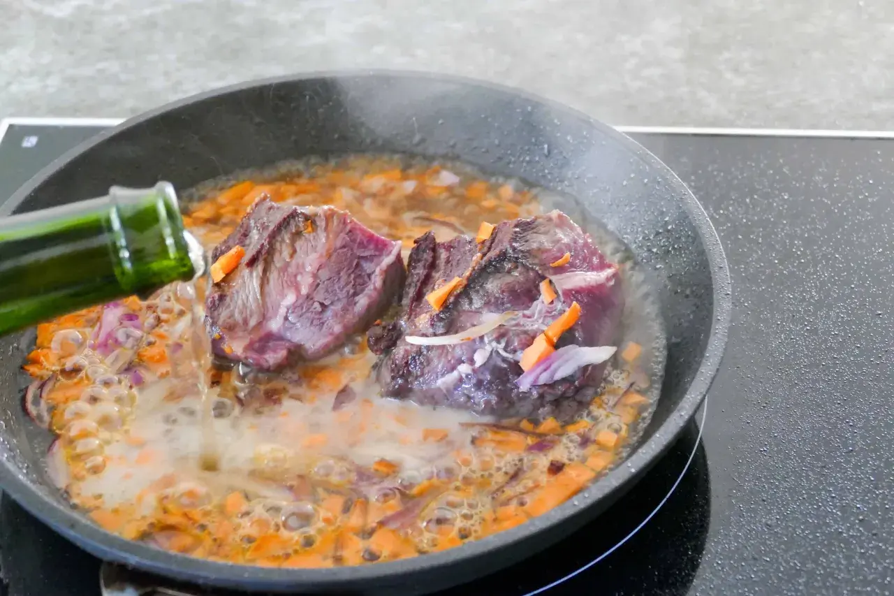 Deux morceaux de joue de bœuf mijotent dans une poêle avec des carottes et des oignons. Un liquide est versé d'une bouteille verte.