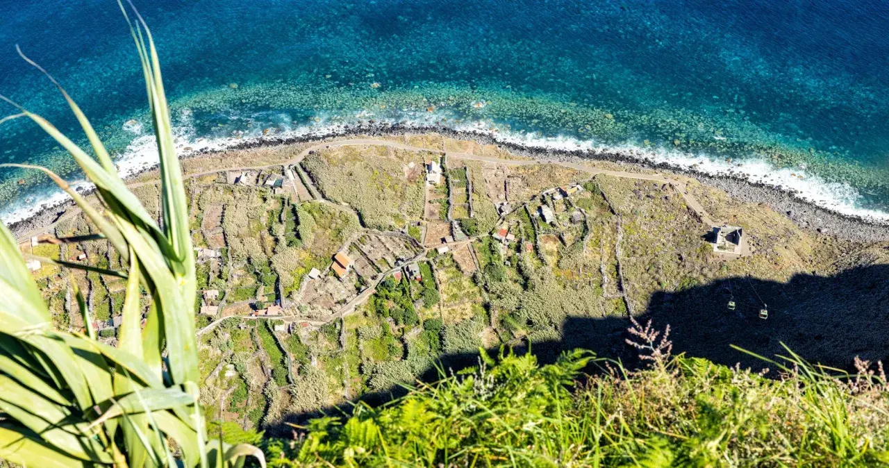 Terraced fields and houses dot the coastline, a glimpse of what to see in Madeira. The turquoise ocean crashes against the rocky shore.