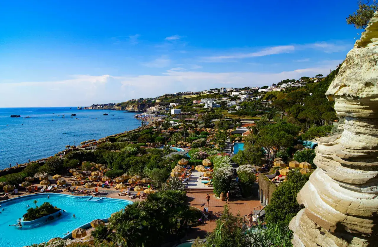 Vista panoramica dell'isola d'Ischia con mare azzurro, piscine, vegetazione lussureggiante e case arroccate sulla collina.