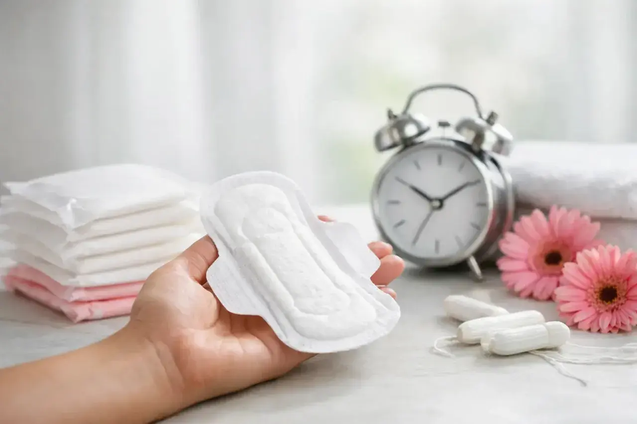 Dłoń trzyma podpaskę, obok stos podpasek, tampony, kwiaty i budzik. Wskazówka: jak podkładać basen sanitarny.