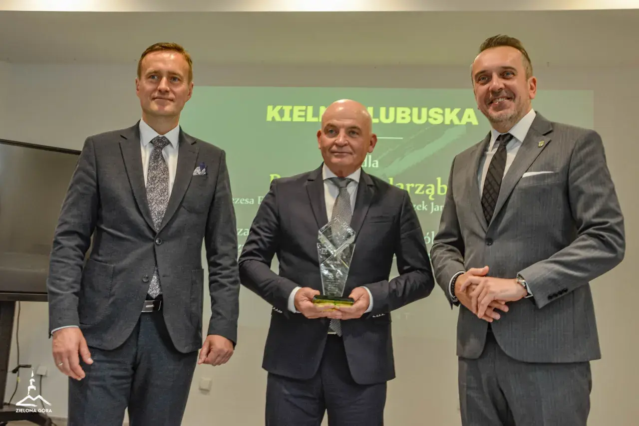 Trzech mężczyzn w garniturach pozuje do zdjęcia, jeden z nich trzyma statuetkę.