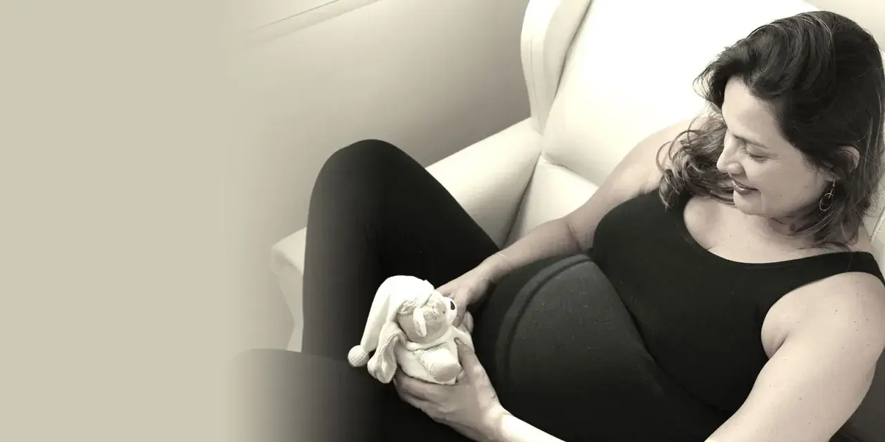 Gravid kvinna i svart klänning håller en nallebjörn. Hon ler och vilar i en stol, kanske under sin sjukskrivning gravid.