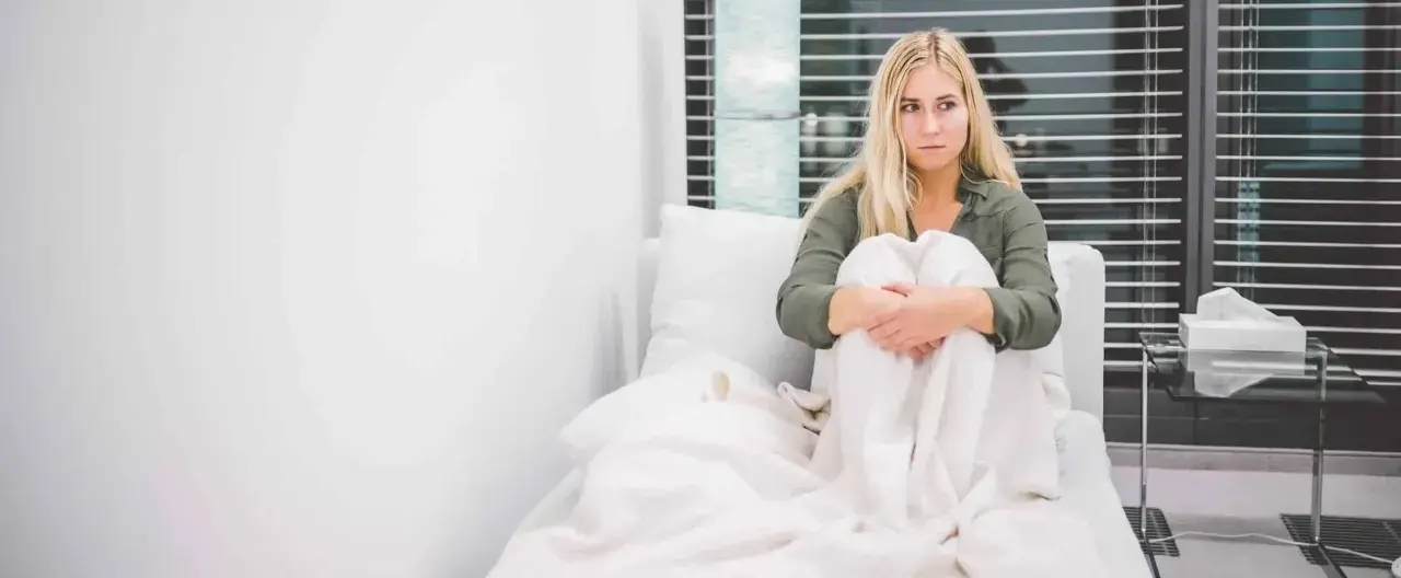 Frau in dissoziativem Stupor, in Decken gehüllt, sitzt auf einem Sofa, Blick leer, Fenster im Hintergrund.