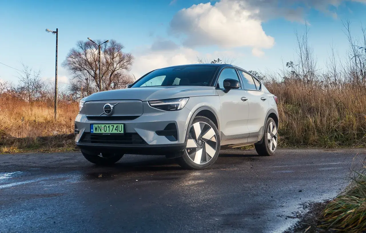 Szary Volvo C40 na mokrej drodze, otoczony suchą trawą i drzewami.