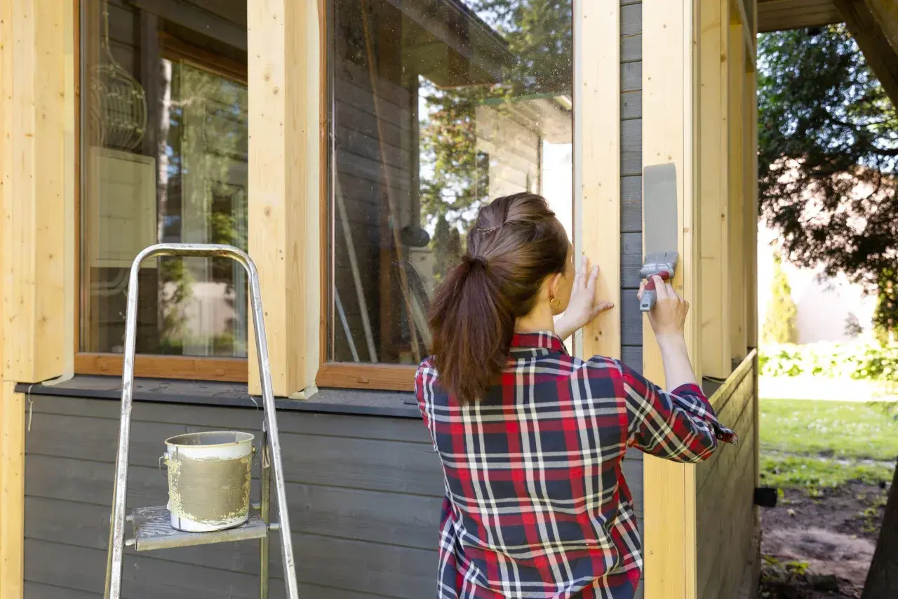 Kvinna målar i solsken en träpanel på ett hus. En stege och en färgburk står bredvid.
