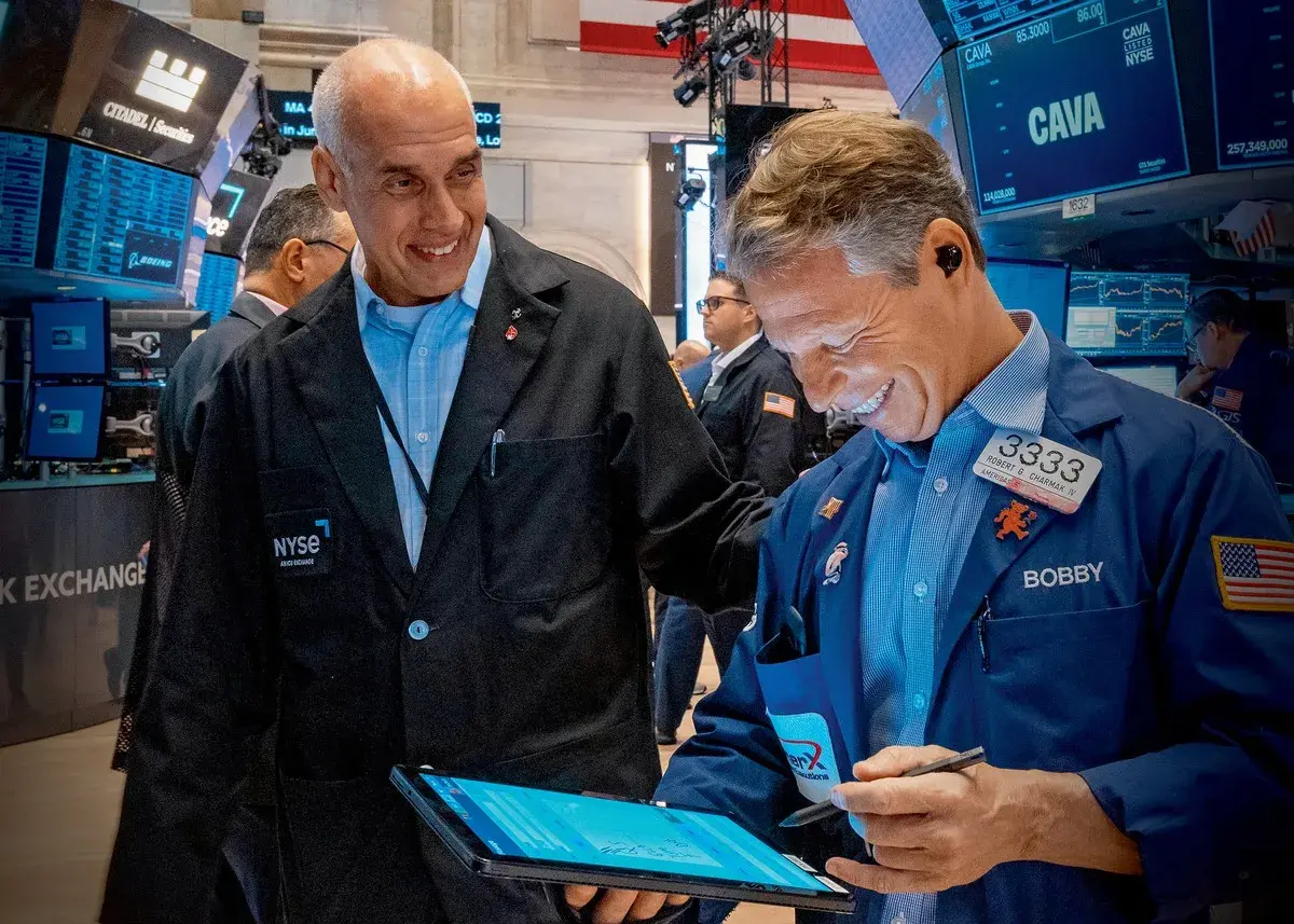 Dwóch mężczyzn w kurtkach NYSE śmieje się, patrząc na tablet. Jeden z nich, Bobby, pokazuje coś na ekranie, a drugi, z napisem "skup akcji własnych" na piersi, uśmiecha się szeroko.