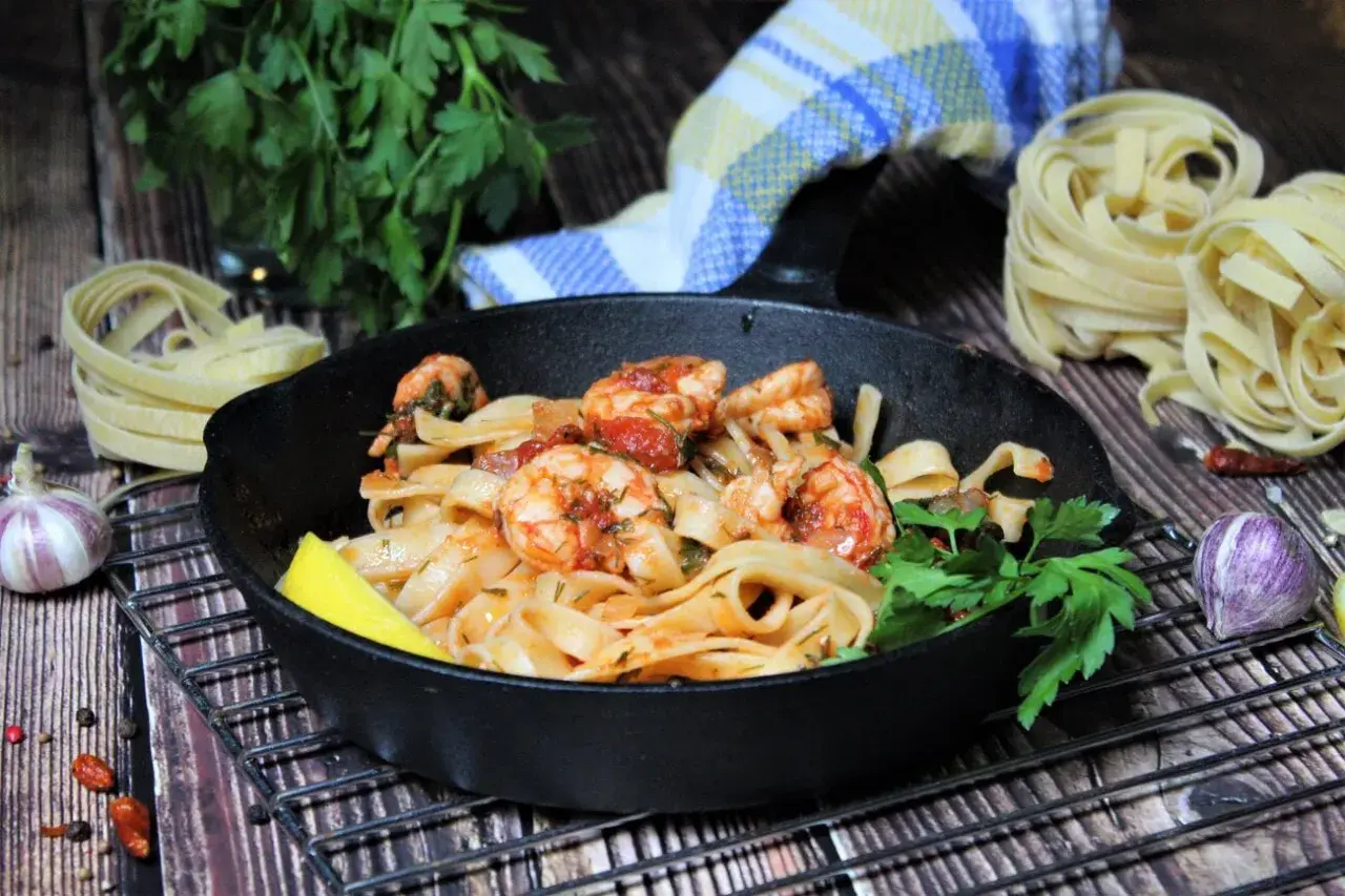 Spaghetti mit Garnelen mediterran in einer Gusseisenpfanne, garniert mit Zitrone und Petersilie. Daneben rohe Pasta, Knoblauch und Chili.