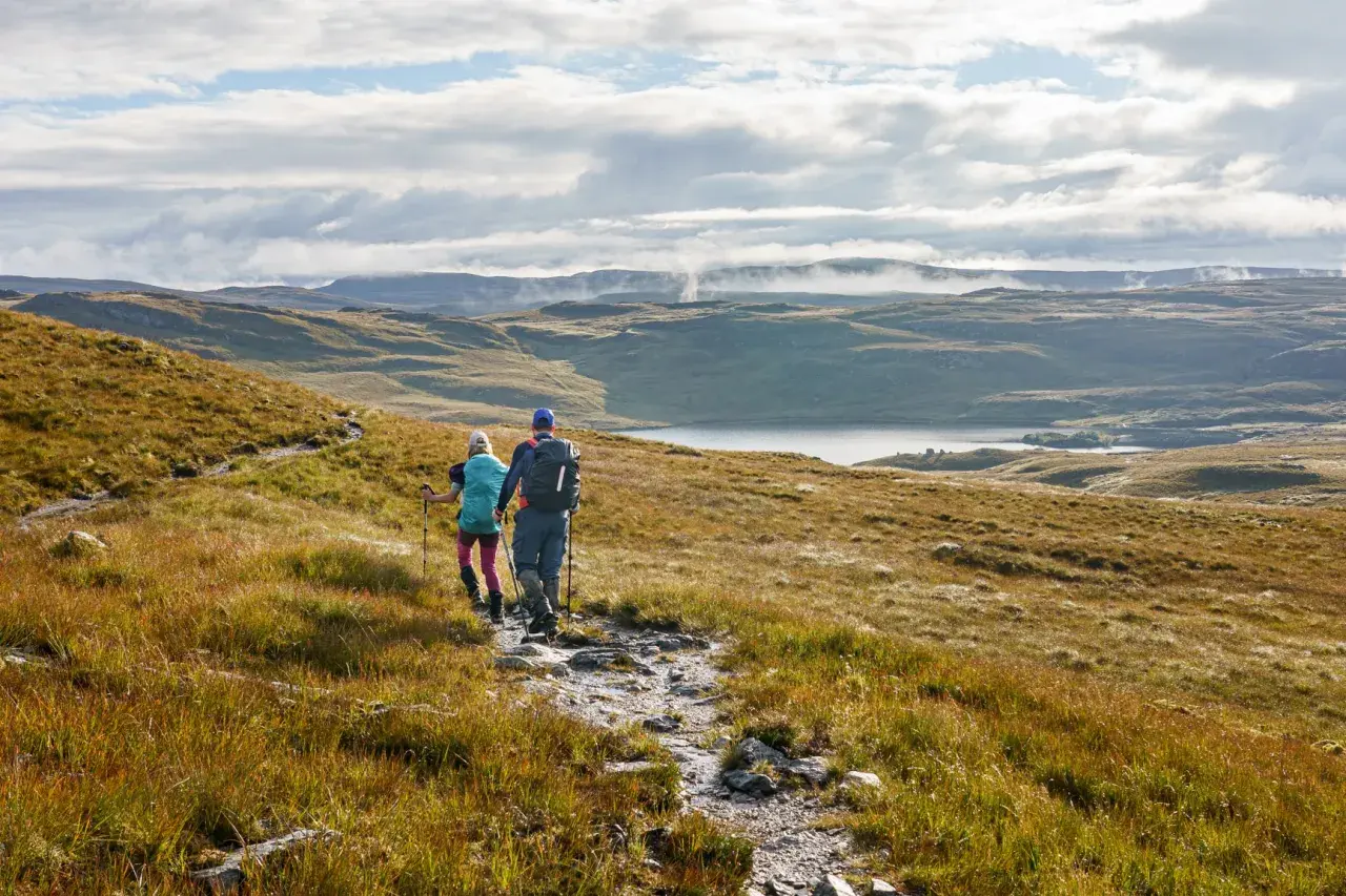 Two hikers trek along the Cape Wrath Trail, a rugged path winding through a vast, misty Scottish landscape with a serene loch in the distance.