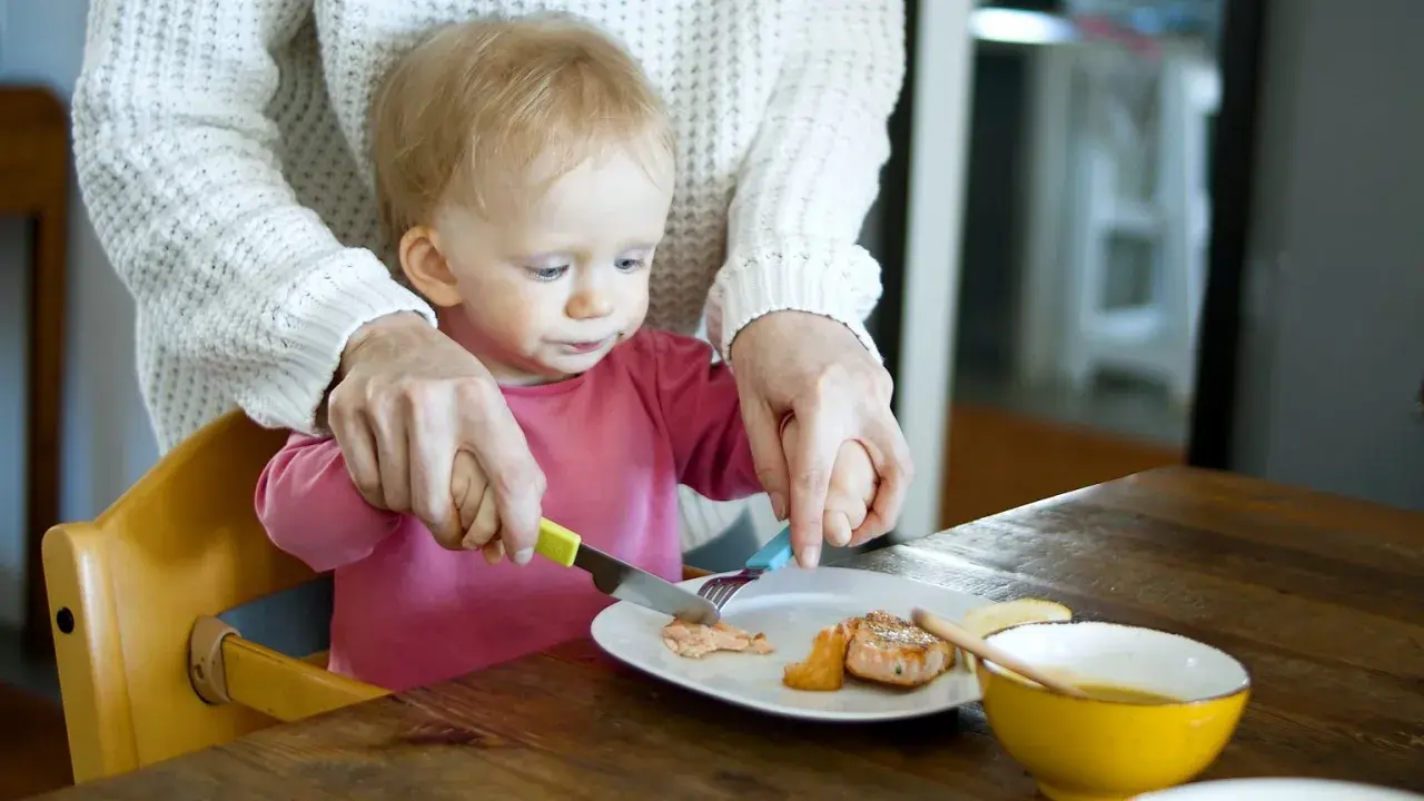 Mama pomaga dziecku kroić rybę na talerzu. To idealne pancakes dla niemowlaka, które uczy się jeść samodzielnie.