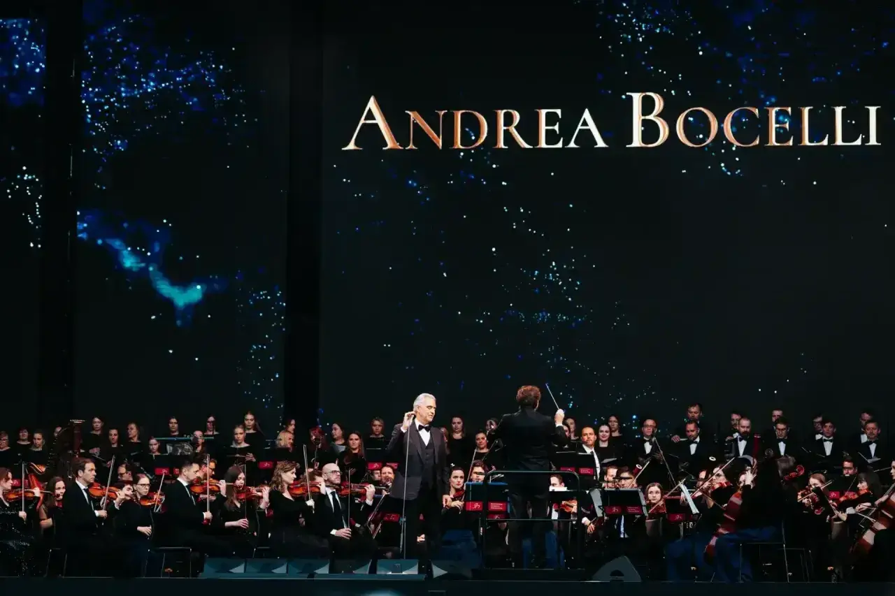 Andrea Bocelli koncert 2026. Wokalista śpiewa z orkiestrą symfoniczną na tle gwieździstego nieba.