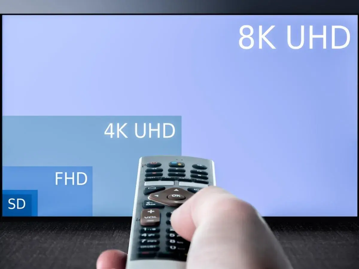 Dłoń trzymająca pilot do telewizora, na ekranie w **hd format** widać porównanie rozdzielczości: 8K UHD, 4K UHD, FHD i SD.