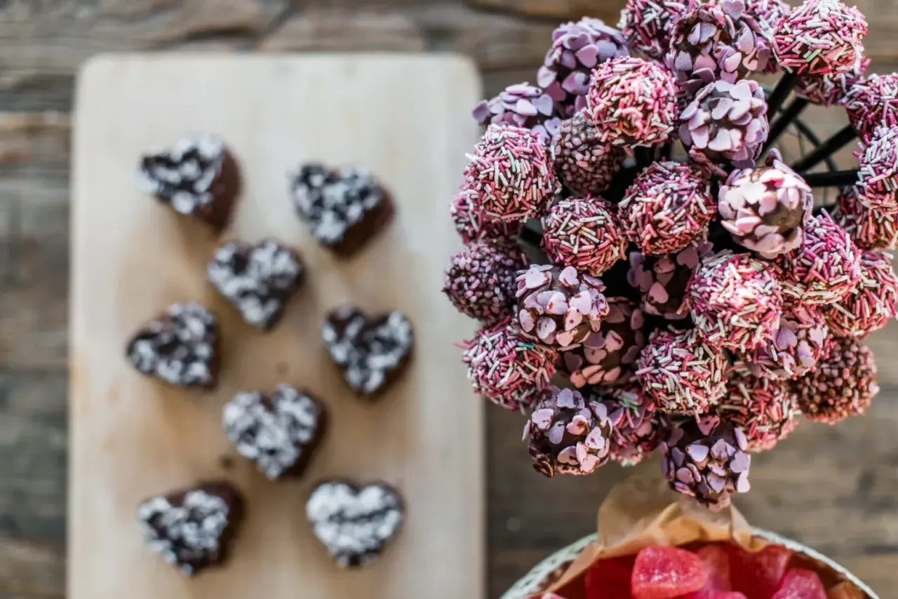 En bukett med rosa och lila cake pops, dekorerade med strössel och hjärtan. Bredvid ligger mörka hjärtformade cake pops.