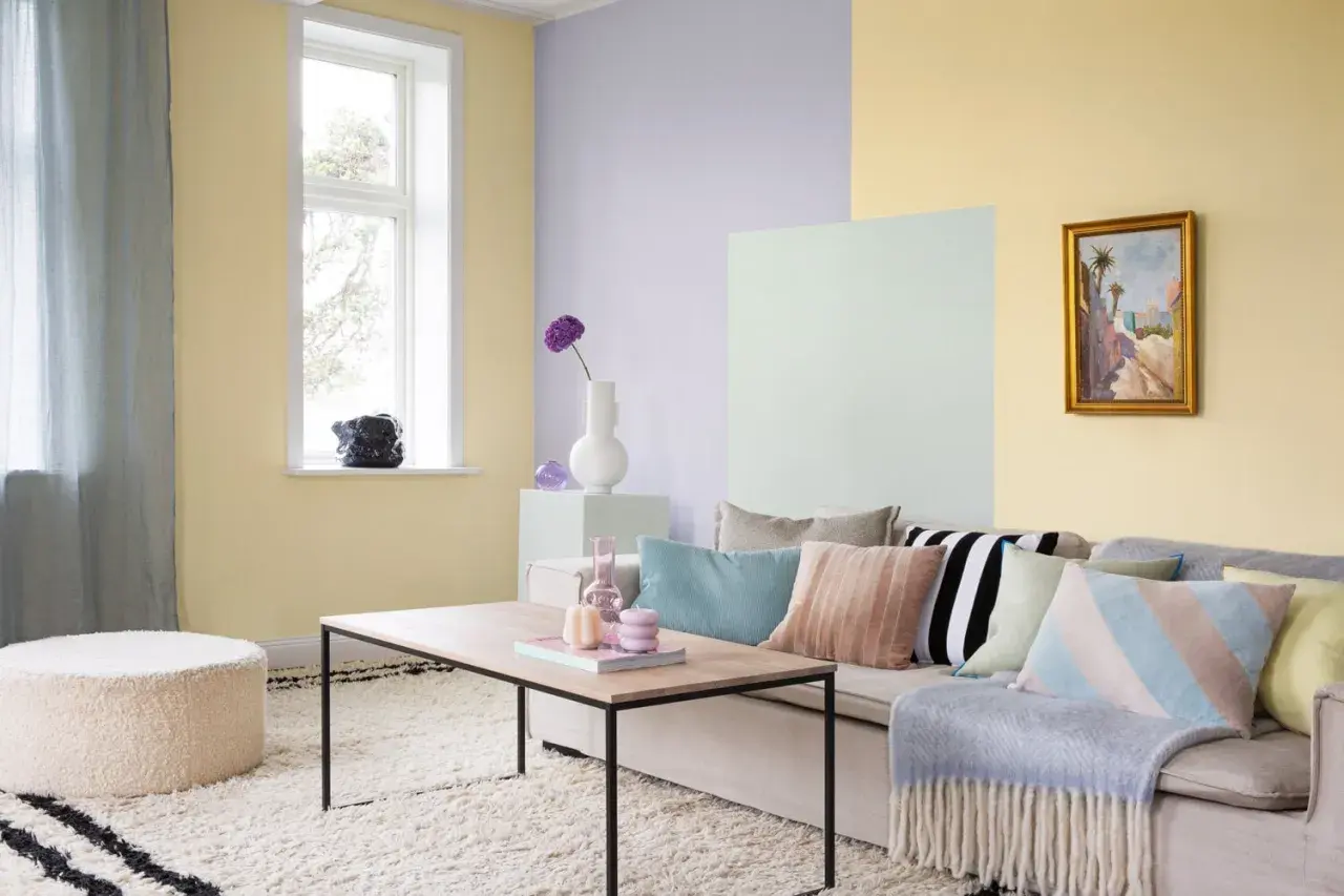 Ett vardagsrum med pastell inredning. Ljusgula, ljuslila och ljusgröna väggar skapar en mjuk atmosfär. En beigefärgad soffa är prydd med kuddar i olika pastellfärger och mönster.