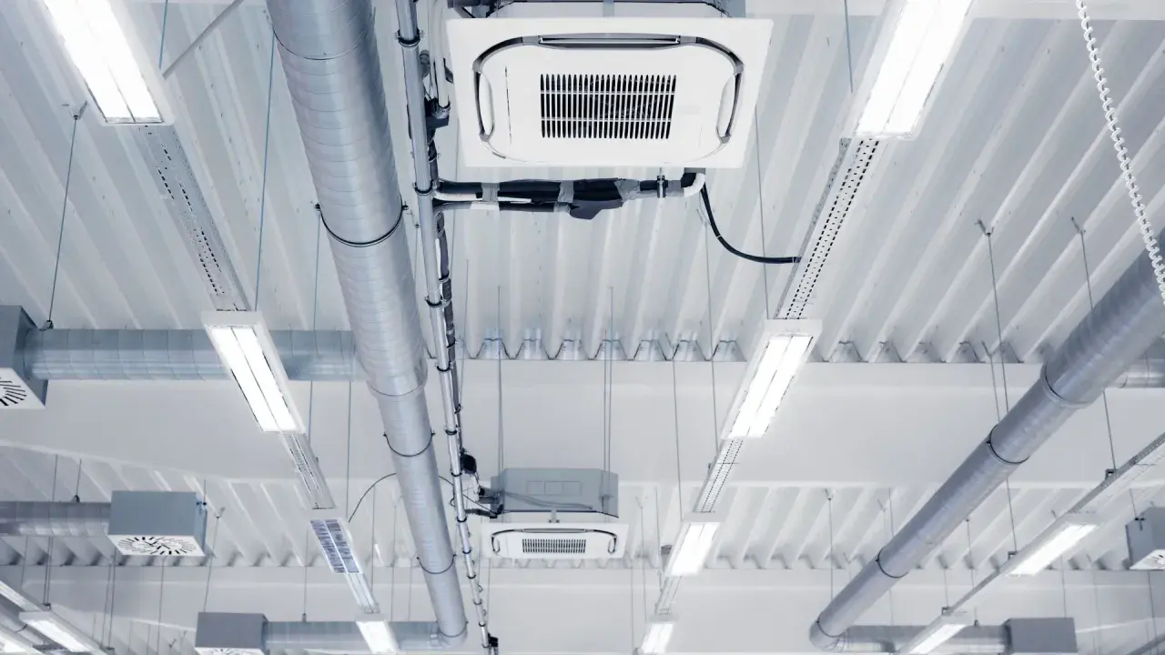 Conductos de ventilación y unidades de aire acondicionado de un sistema VRV, iluminados por luces fluorescentes en un techo industrial.