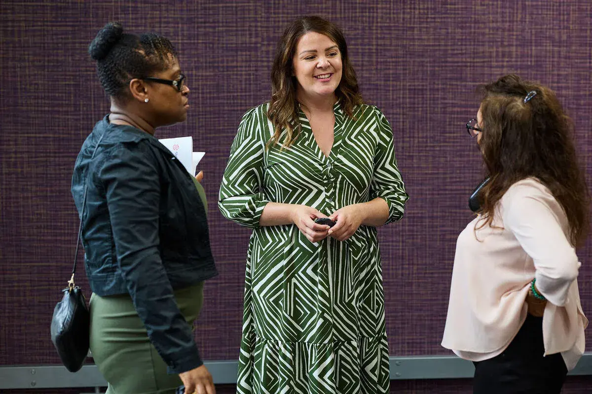 Three women in conversation, one in a green patterned dress, discussing strategies for driving performance.