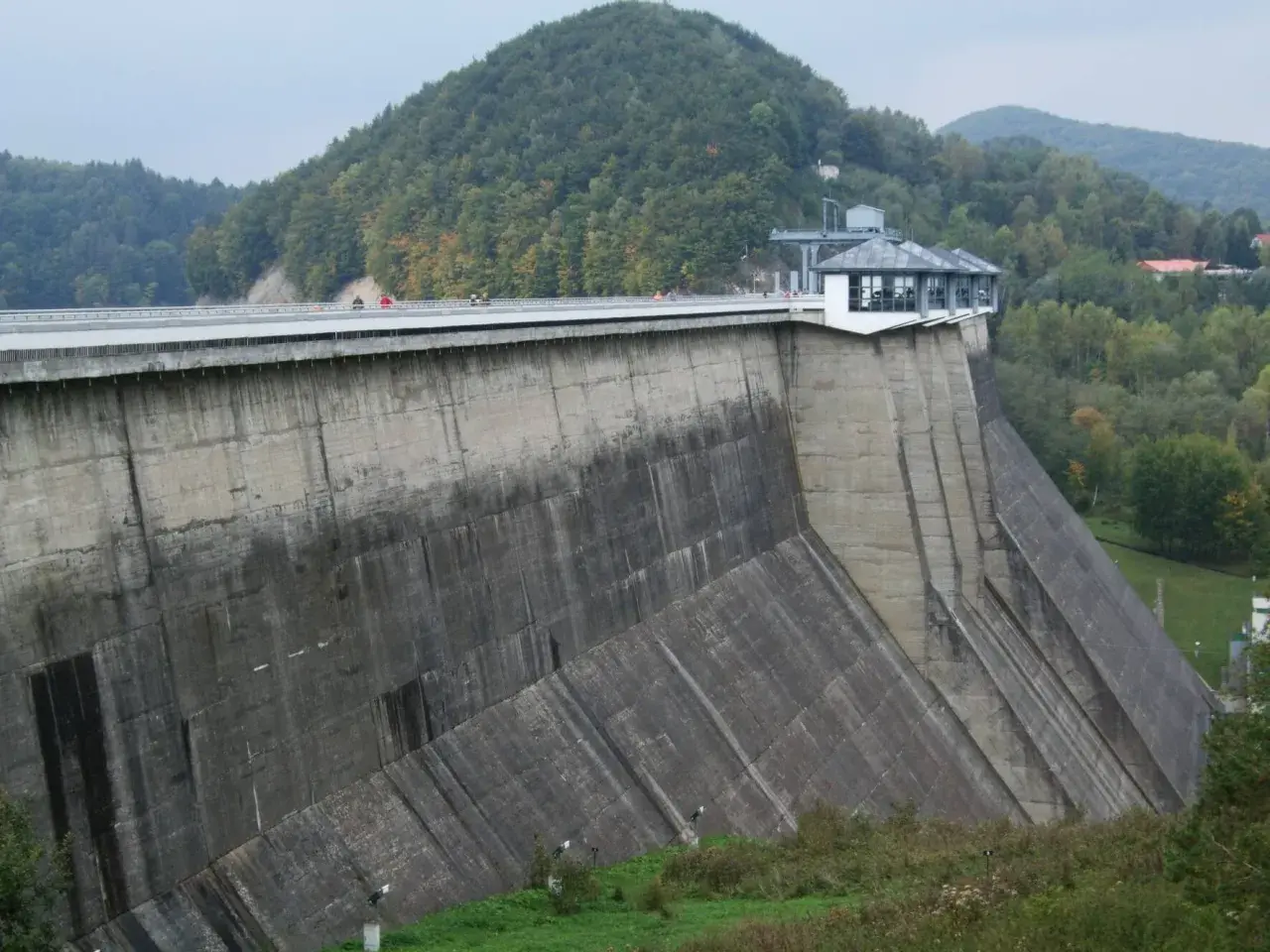 Ogromna zapora na sanie góruje nad zielonymi wzgórzami. Na jej szczycie widać ludzi spacerujących po moście.