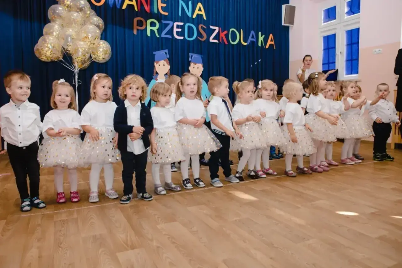 Dzieci w strojach galowych stoją w rzędzie na tle niebieskiej kurtyny z napisem "Pasowanie na przedszkolaka".