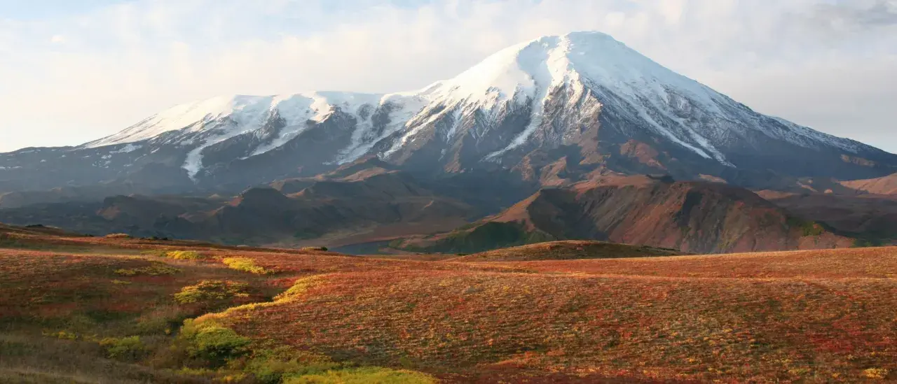 Majestätischer schneebedeckter Vulkan in einer weiten russischen Landschaft mit herbstlich gefärbten Hügeln im Vordergrund.