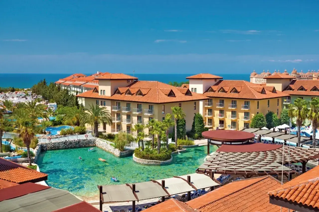 Ein luxuriöser Urlaub im Club Grand Aqua: Sonnige Gebäude mit roten Ziegeldächern, Palmen und ein türkisfarbener Pool mit Badegästen.