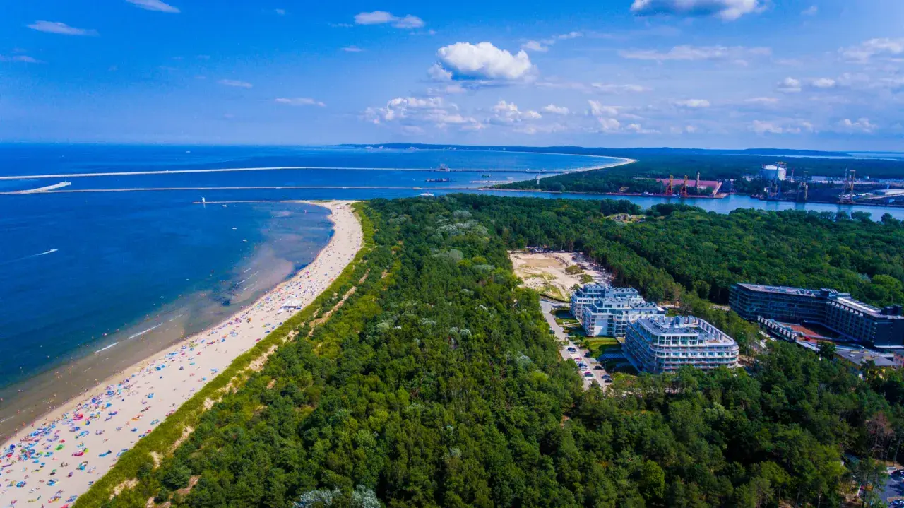 Plaża w Świnoujściu z lotu ptaka. Tłumy plażowiczów, turkusowe morze i zielony las otaczający nowoczesne apartamenty.