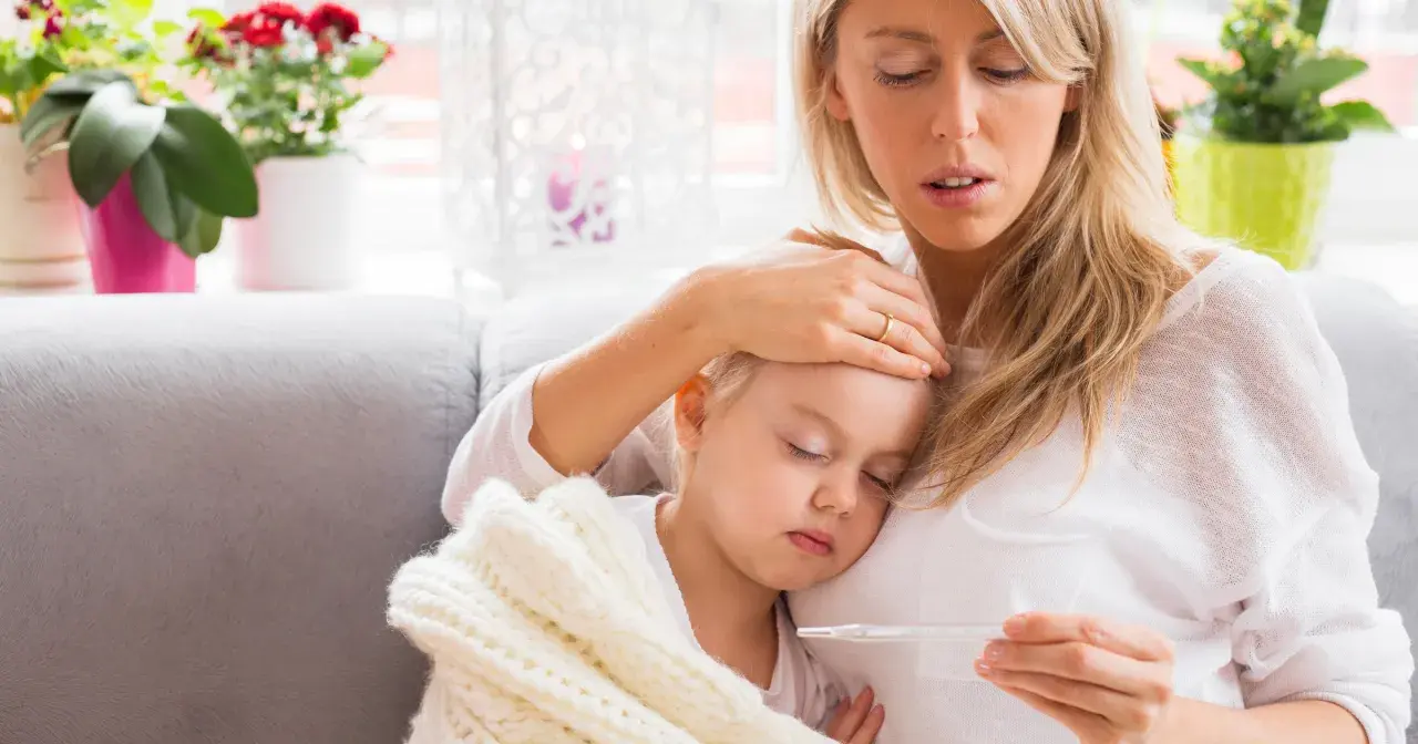 Zmartwiona mama sprawdza temperaturę chorej córki, która śpi otulona kocykiem. Myśli o podaniu Fervex dla dzieci.