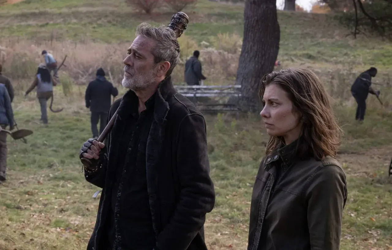 Maggie et Negan, le regard lointain, se tiennent côte à côte dans un paysage automnal. D'autres survivants travaillent en arrière-plan.