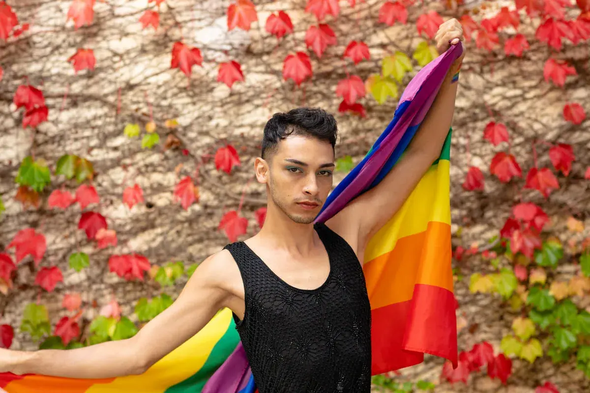 Un giovane con la bandiera arcobaleno, simbolo del **queer significato** di libertà e identità, si staglia contro un muro fiorito.