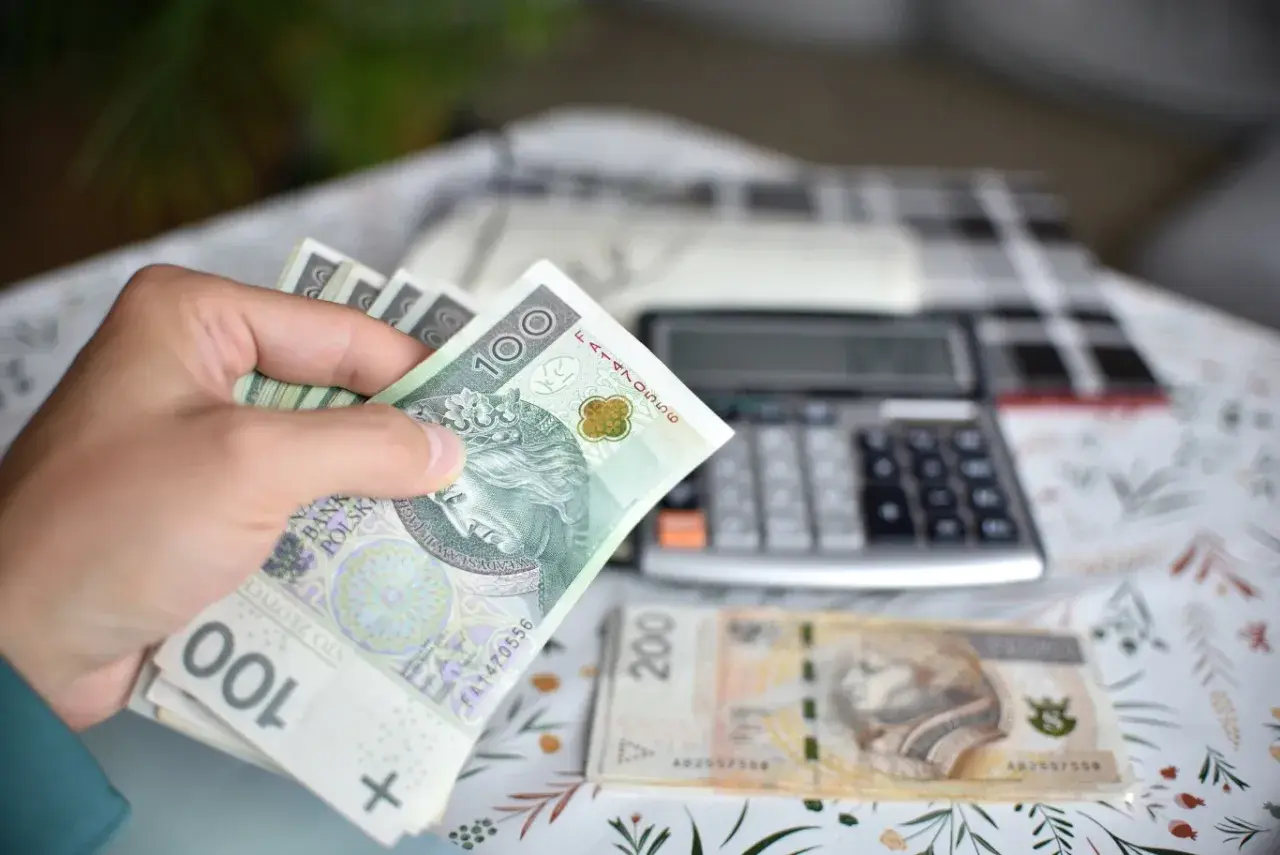 Ręka trzyma banknoty 100 zł, obok leży 200 zł i kalkulator. Obliczenia ekwiwalentu za urlop.