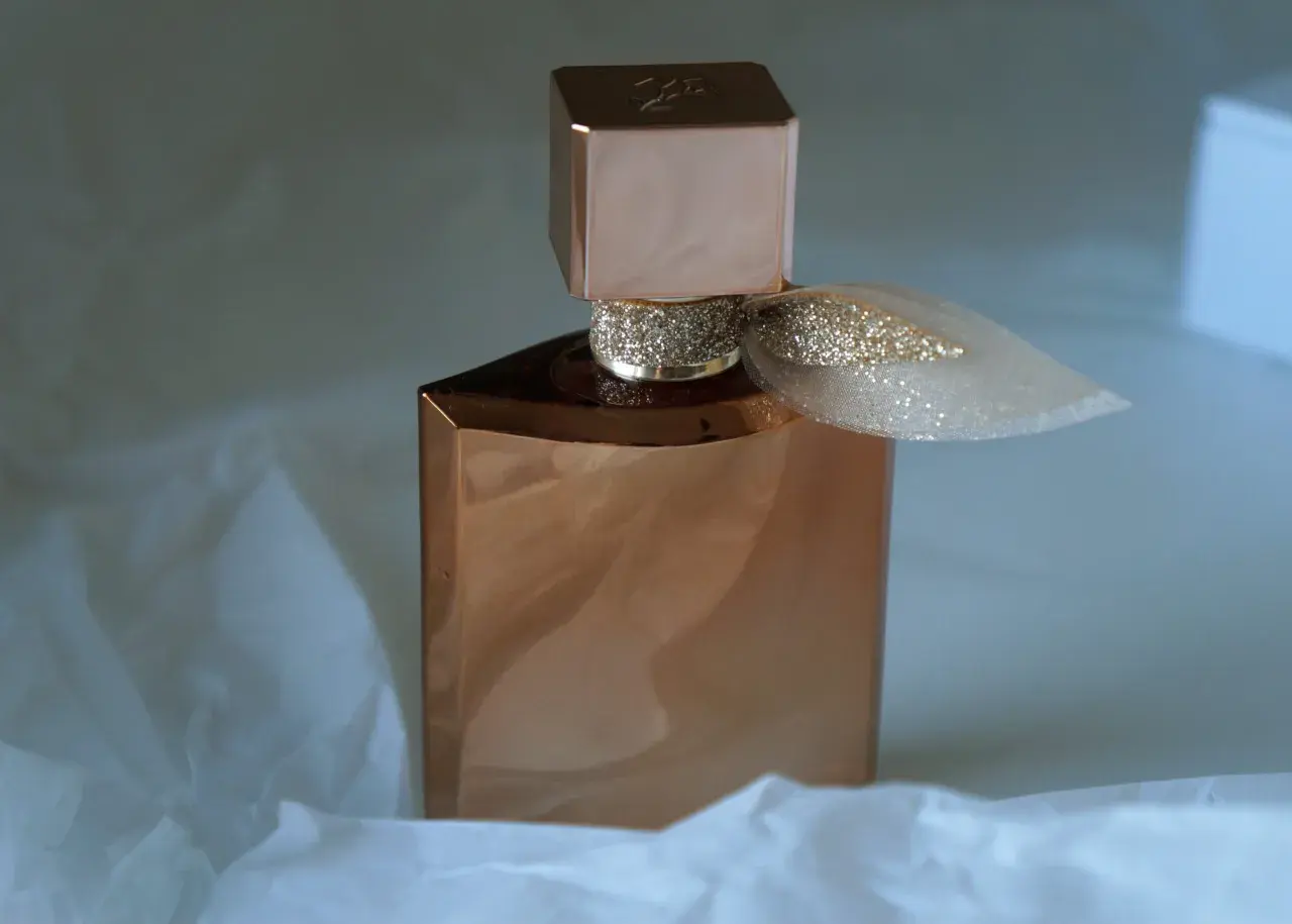 Elegancka butelka perfum Lancome Eclat w kolorze miedzi, ozdobiona brokatową wstążką, otulona delikatnym białym papierem.