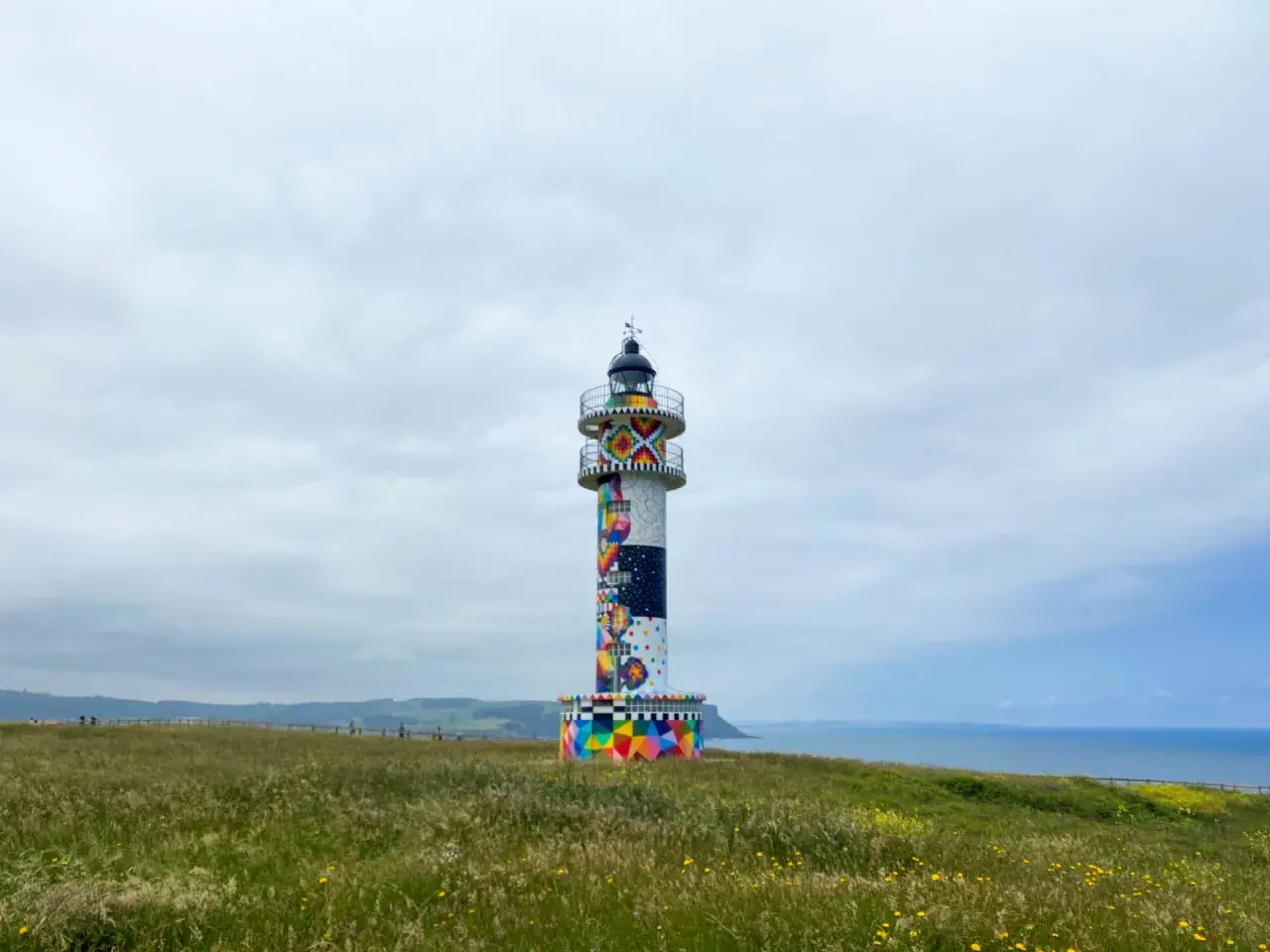 Faro colorido en el cabo de Ajo, con un paisaje verde y el mar al fondo.